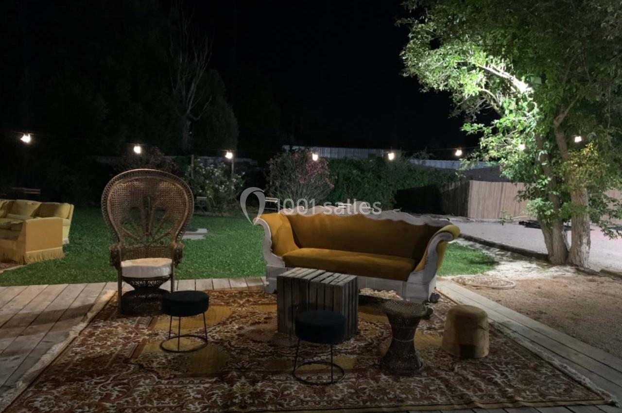 Espace extérieur nocturne avec canapé, fauteuils et tapis éclairés par des guirlandes lumineuses, entouré de verdure.