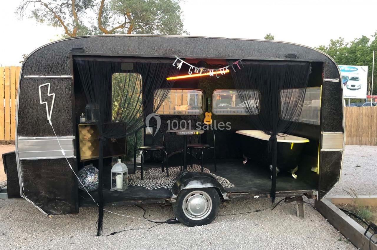 Caravane aménagée avec une baignoire, une guitare et des décorations, ouverte sur un espace extérieur gravillonné.