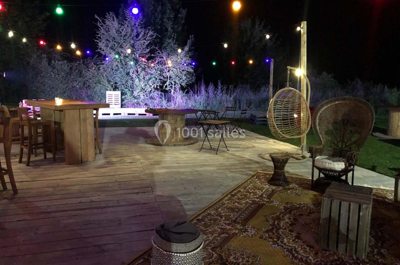 Terrasse en bois éclairée par des guirlandes lumineuses colorées, avec mobilier en rotin et tables en extérieur.