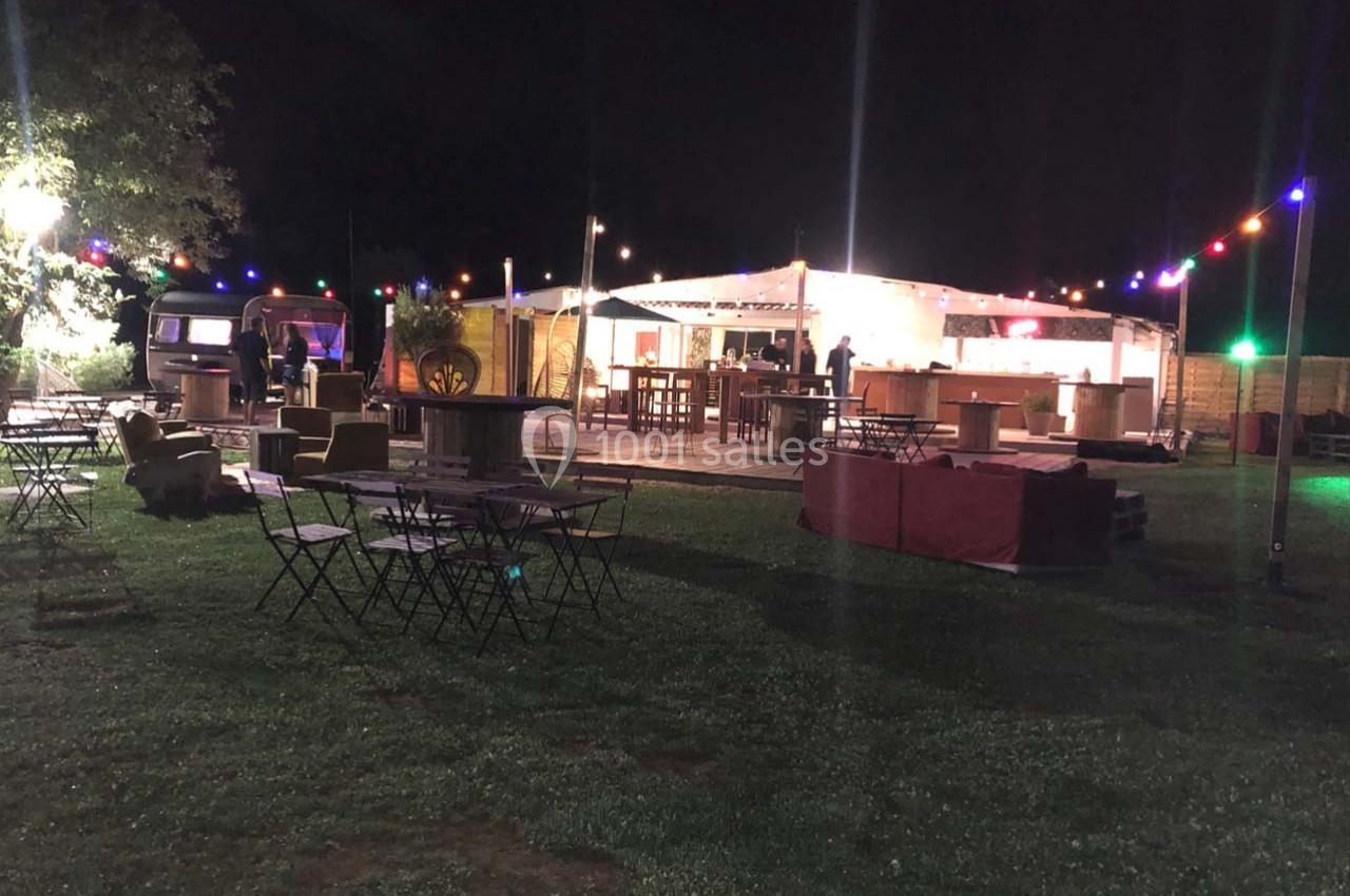 Espace extérieur nocturne avec tables, chaises et guirlandes lumineuses, devant un bar éclairé sous une tente.