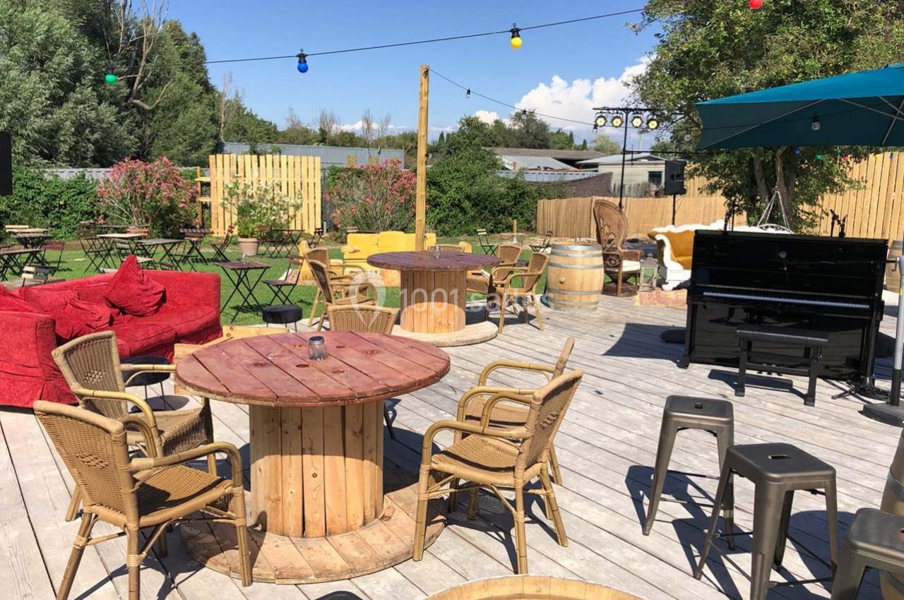 Terrasse en bois avec tables et chaises en plein air, décorée de guirlandes lumineuses et entourée de verdure.