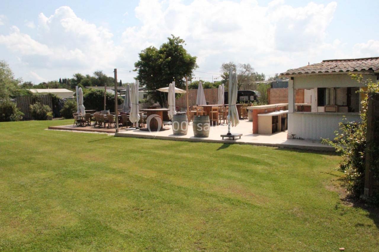Terrasse extérieure avec tables, chaises, parasols et un bar en bois, entourée d'une pelouse et d'arbres.