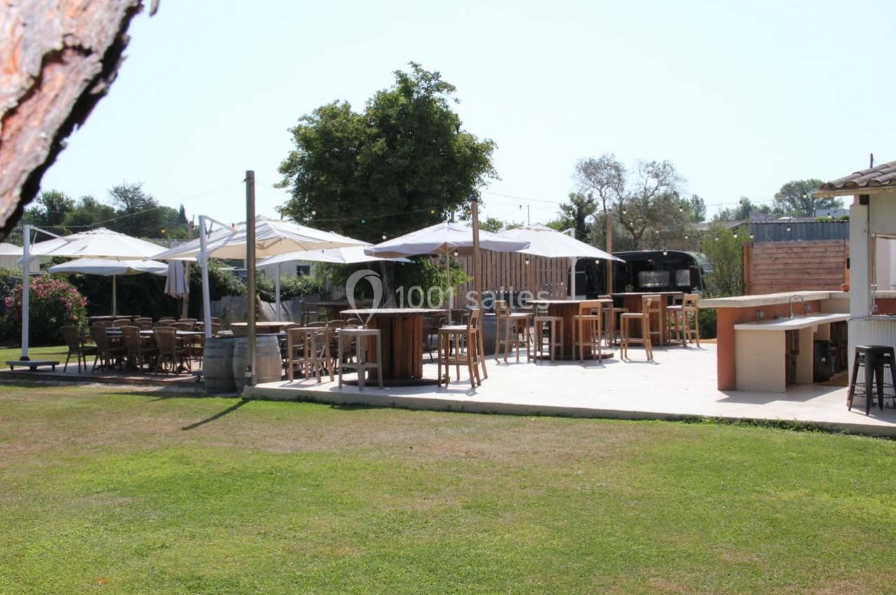 Terrasse extérieure avec tables, chaises et parasols, entourée de verdure et d'arbres sous un ciel dégagé.