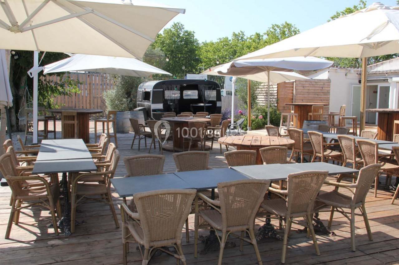Terrasse en bois aménagée avec des tables, chaises en rotin et parasols, entourée de verdure et d'un food truck noir.
