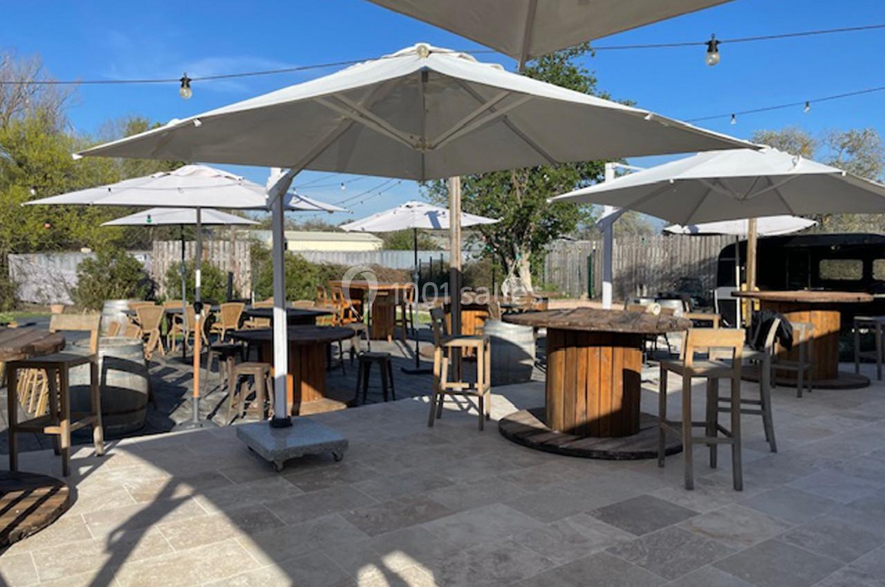 Terrasse extérieure avec tables hautes en bois, chaises et parasols blancs, entourée de verdure sous un ciel dégagé.