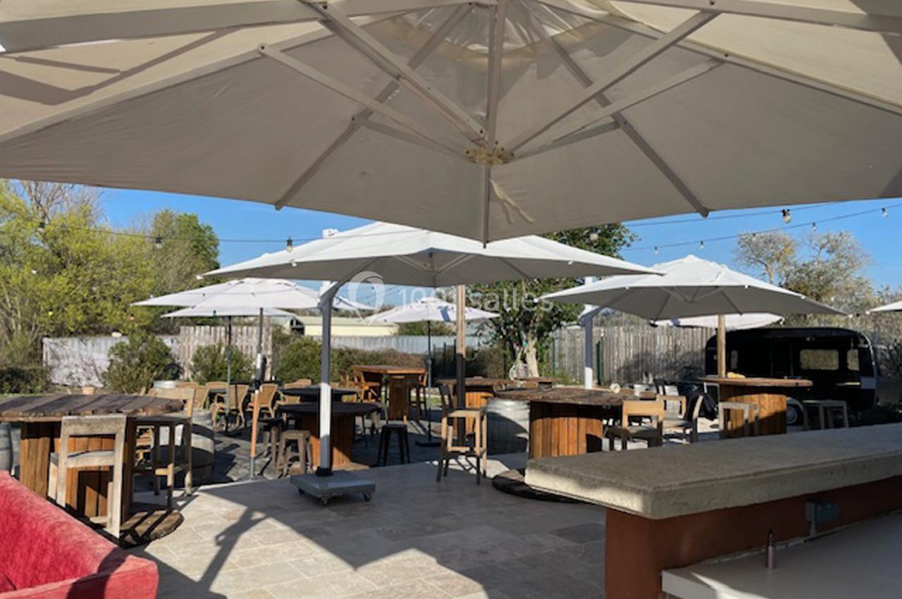Terrasse extérieure avec tables en bois et parasols blancs, entourée de végétation sous un ciel dégagé.