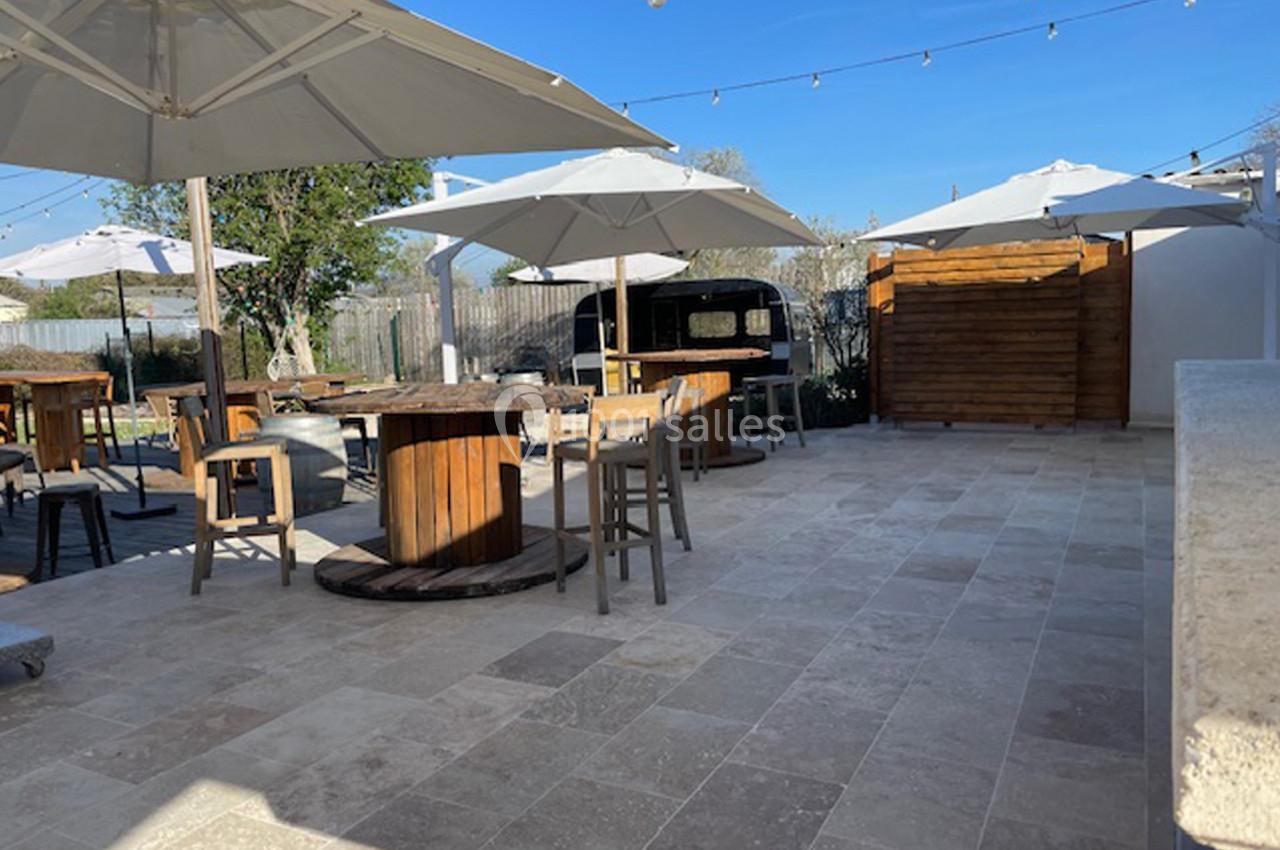 Terrasse extérieure avec tables en bois, chaises, parasols blancs et un comptoir en bois sous un ciel dégagé.