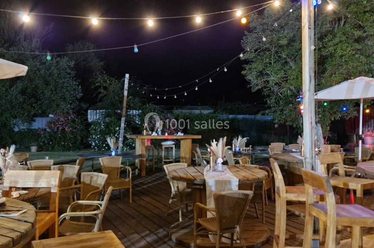 Terrasse en bois éclairée par des guirlandes lumineuses, avec des tables et chaises en bois, entourée de végétation.