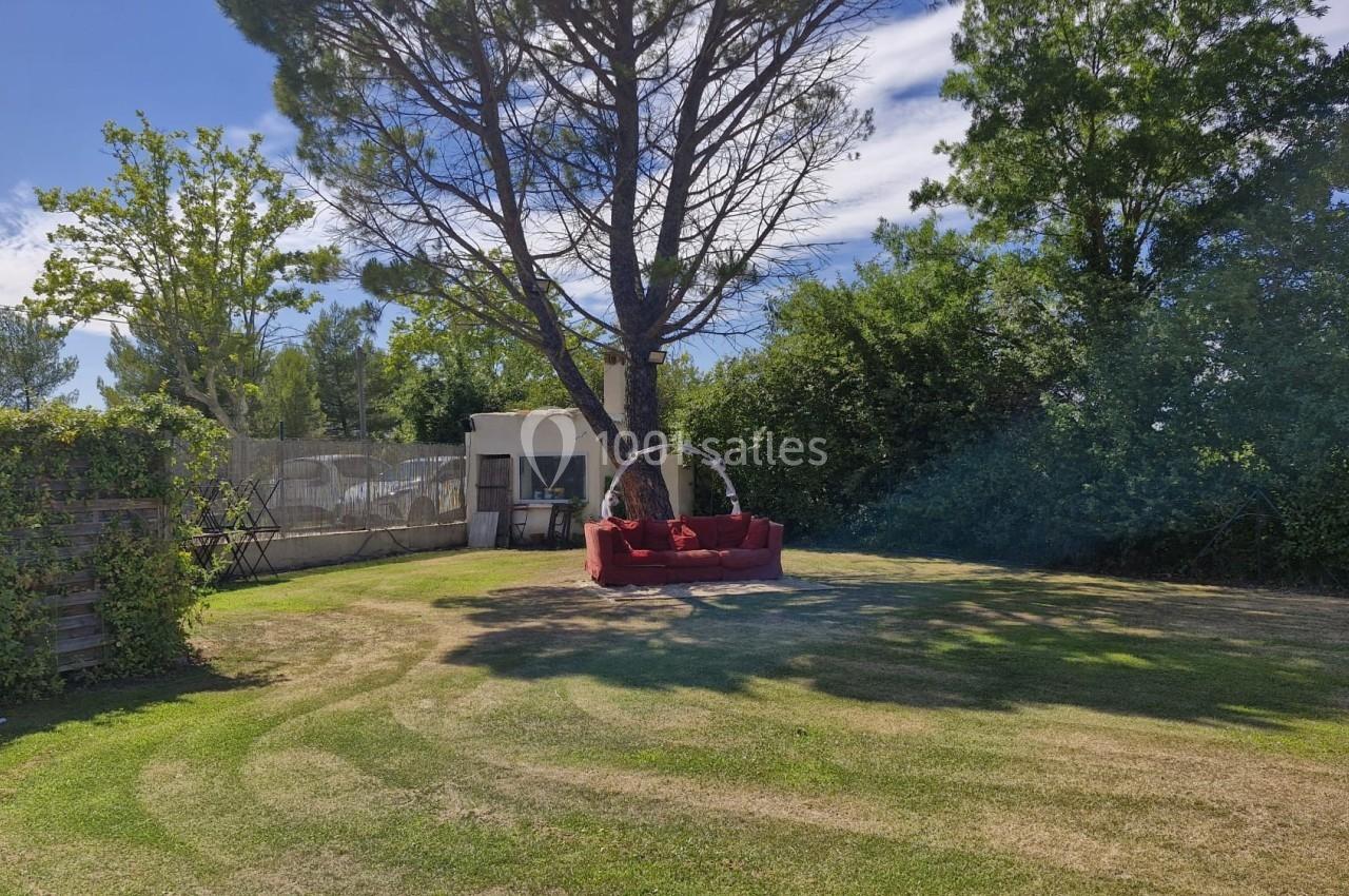 Un canapé rouge est installé sous un grand arbre dans un jardin ensoleillé entouré de végétation.