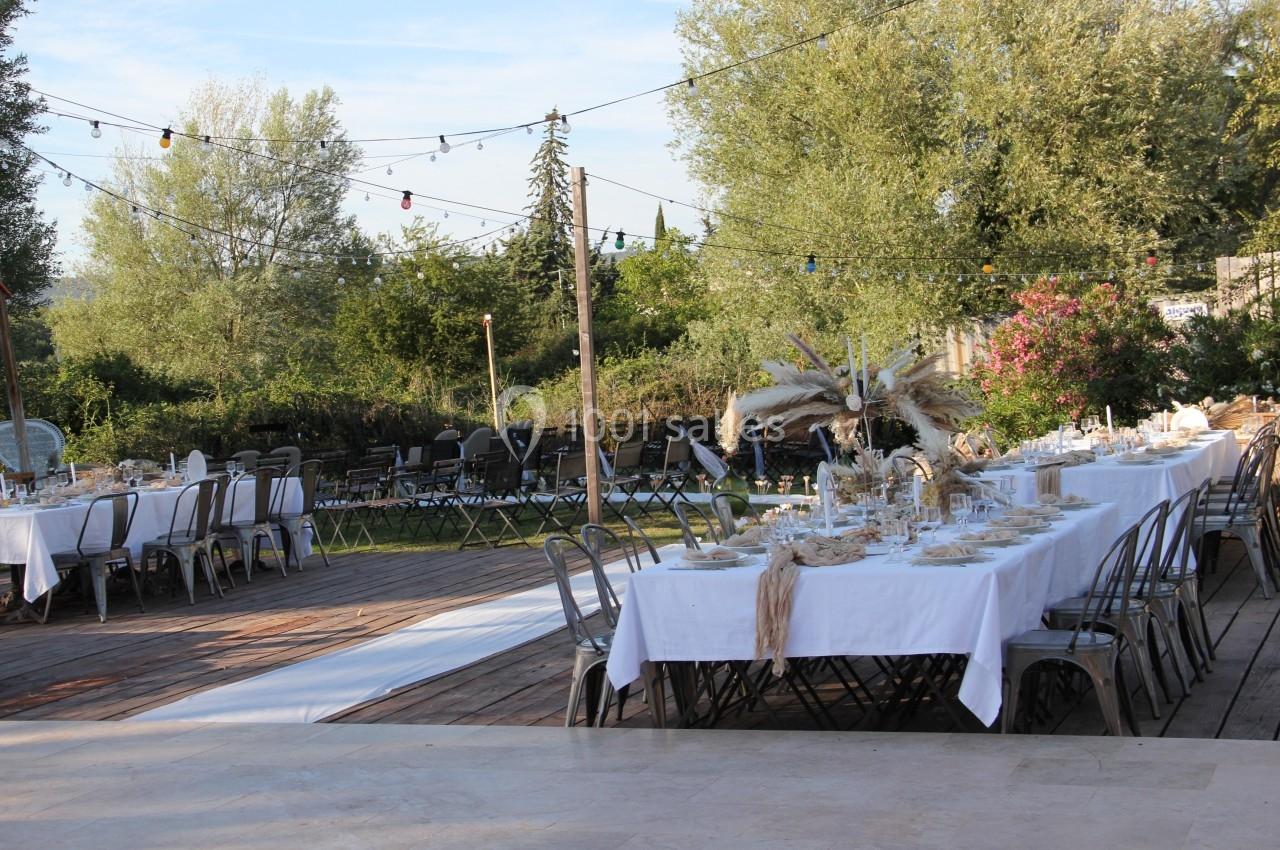 Tables dressées en extérieur avec nappes blanches, chaises métalliques et décorations naturelles sous des guirlandes…