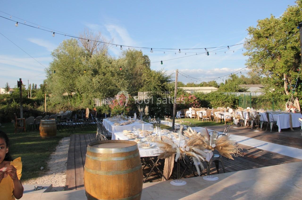 Tables décorées pour un événement en plein air avec guirlandes lumineuses, barriques en bois et végétation en arrière-plan.