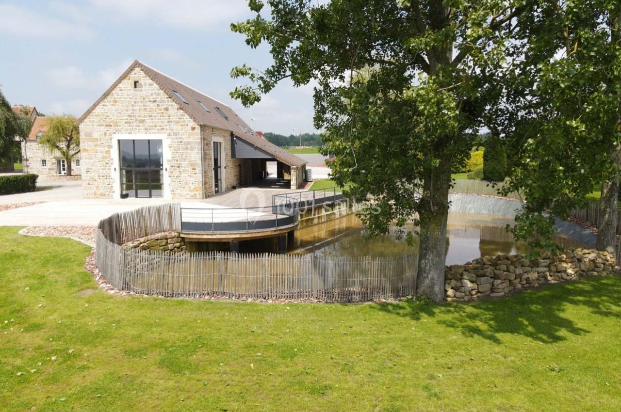 Maison en pierre avec terrasse surplombant un petit étang entouré d'une clôture en bois, dans un cadre verdoyant.