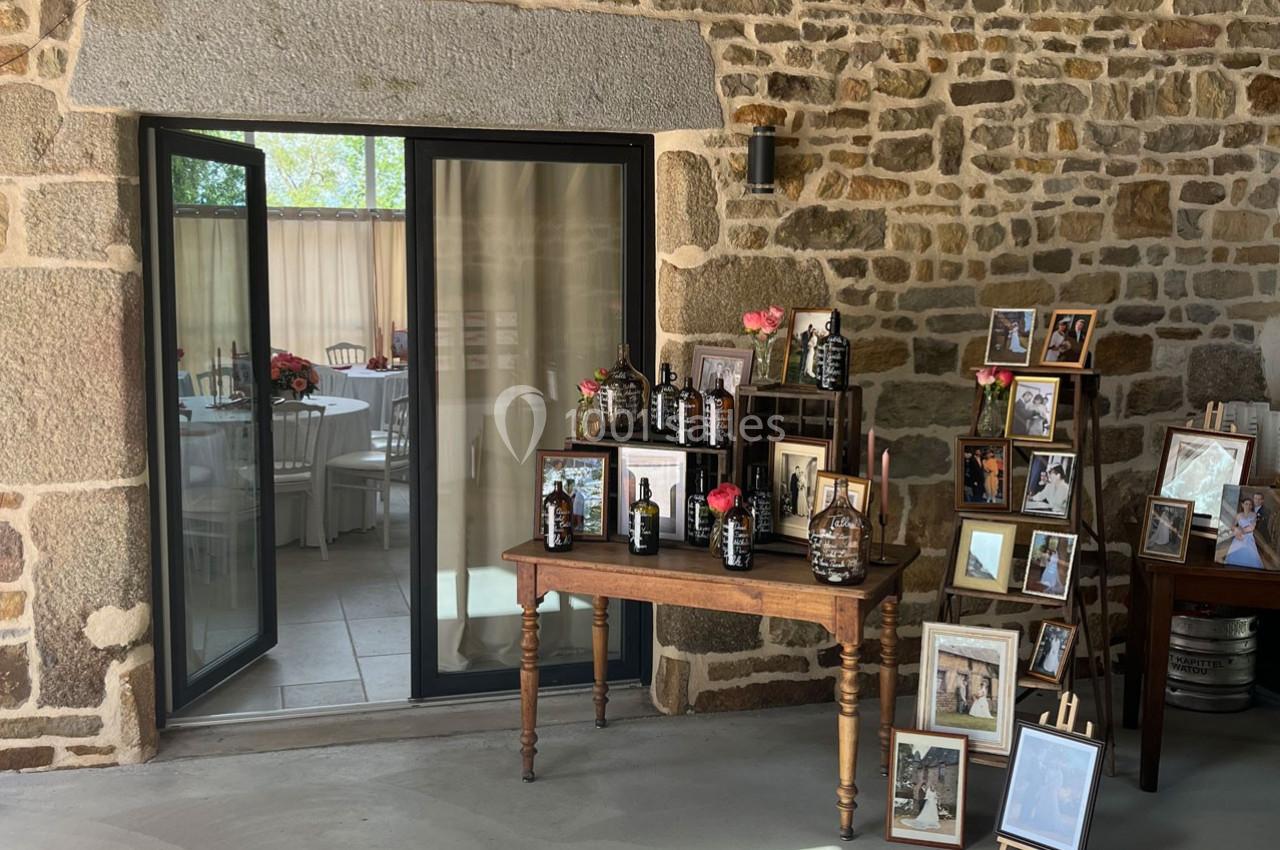 Table en bois avec des bouteilles décoratives et des fleurs, entourée de cadres photo, devant un mur en pierre et une porte…