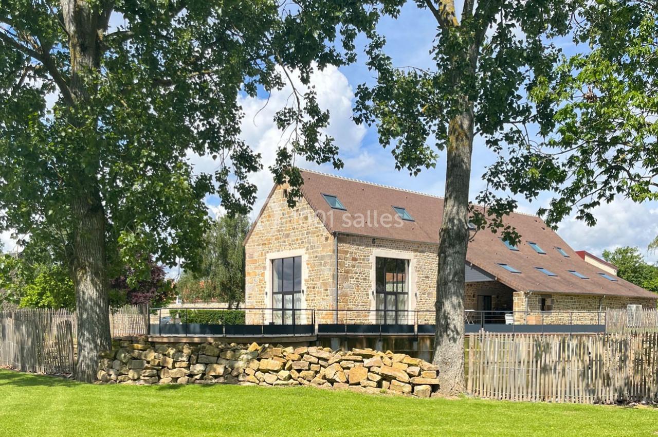 Maison en pierre avec toit en pente, entourée d'arbres et d'une pelouse, sous un ciel partiellement nuageux.