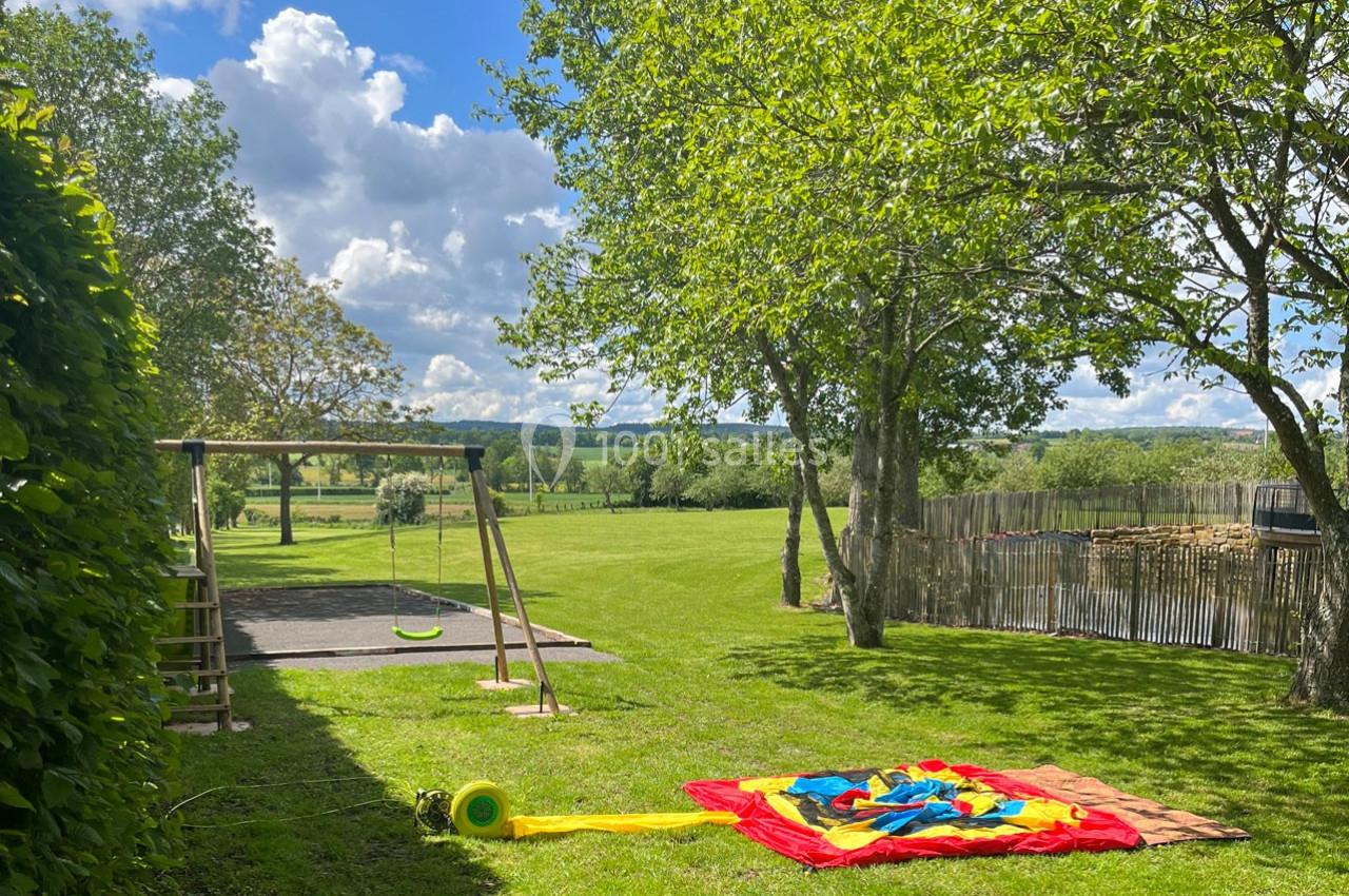 Jardin verdoyant avec balançoire, arbres, pelouse et un tapis coloré posé au sol sous un ciel partiellement nuageux.