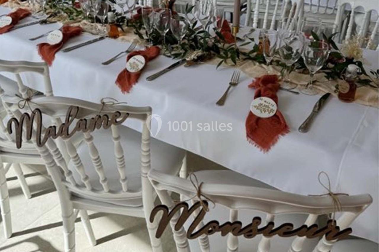 Table de réception décorée avec des couverts, serviettes rouges, feuillage et chaises marquées ’Madame’ et ’Monsieur’.