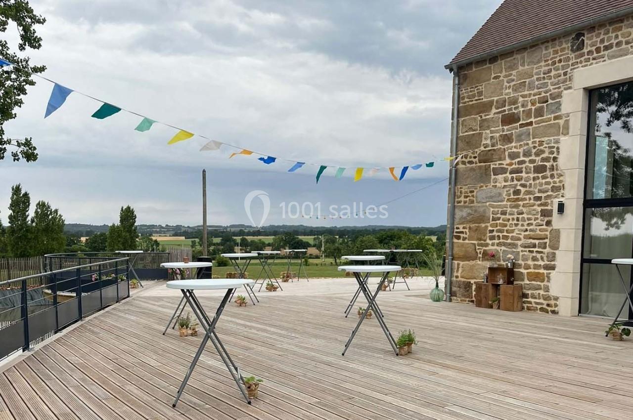 Terrasse en bois avec tables hautes, guirlandes colorées et vue sur un paysage champêtre sous un ciel nuageux.