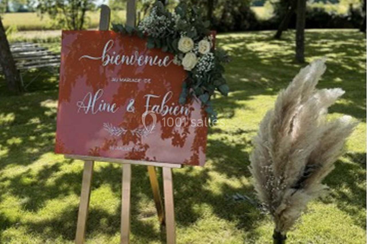 Panneau de bienvenue rouge sur un chevalet, décoré de fleurs blanches et de pampas, posé sur une pelouse ensoleillée.