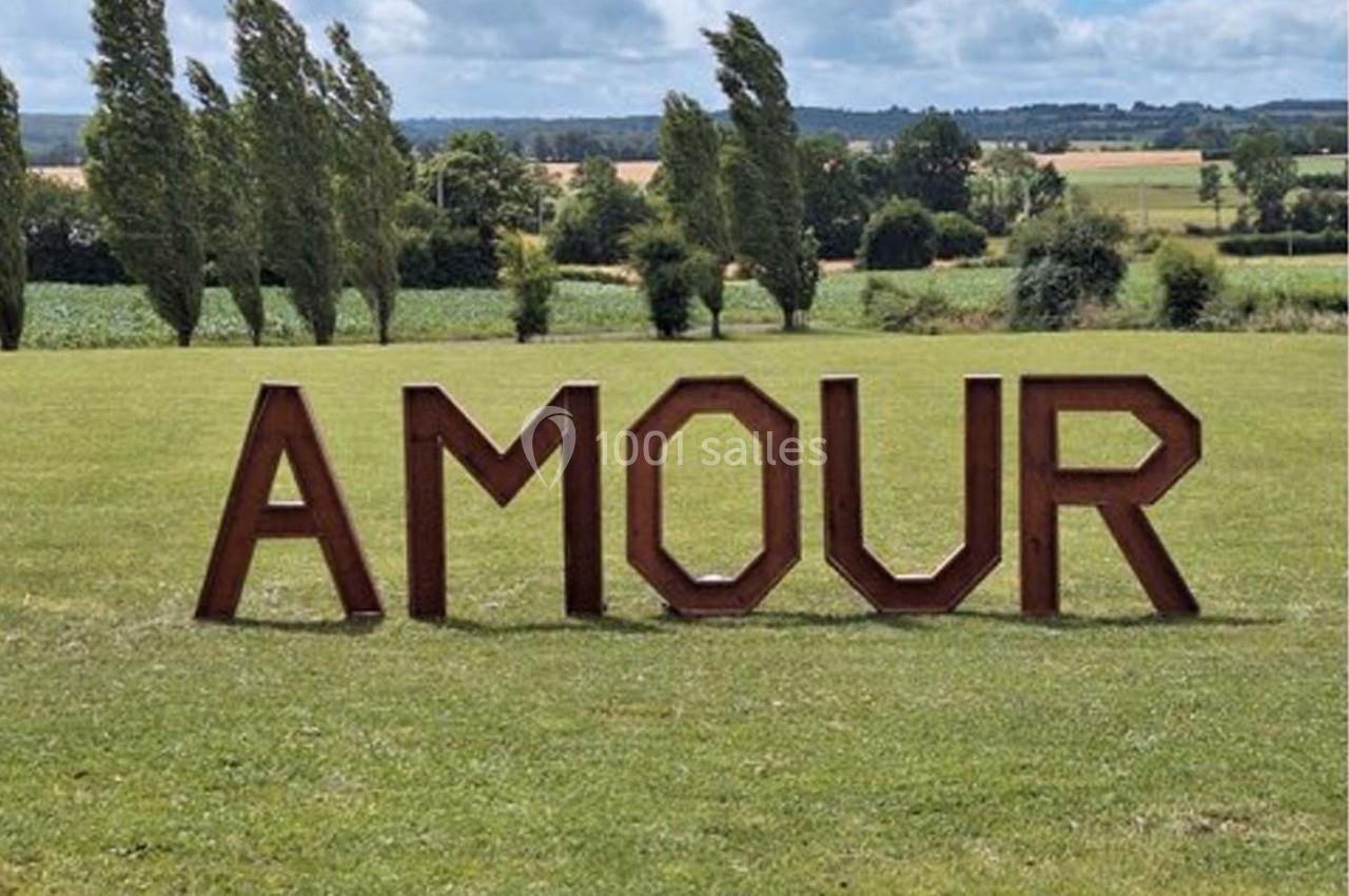 Lettres en bois formant le mot ’AMOUR’ disposées sur une pelouse, avec des arbres et un paysage rural en arrière-plan.