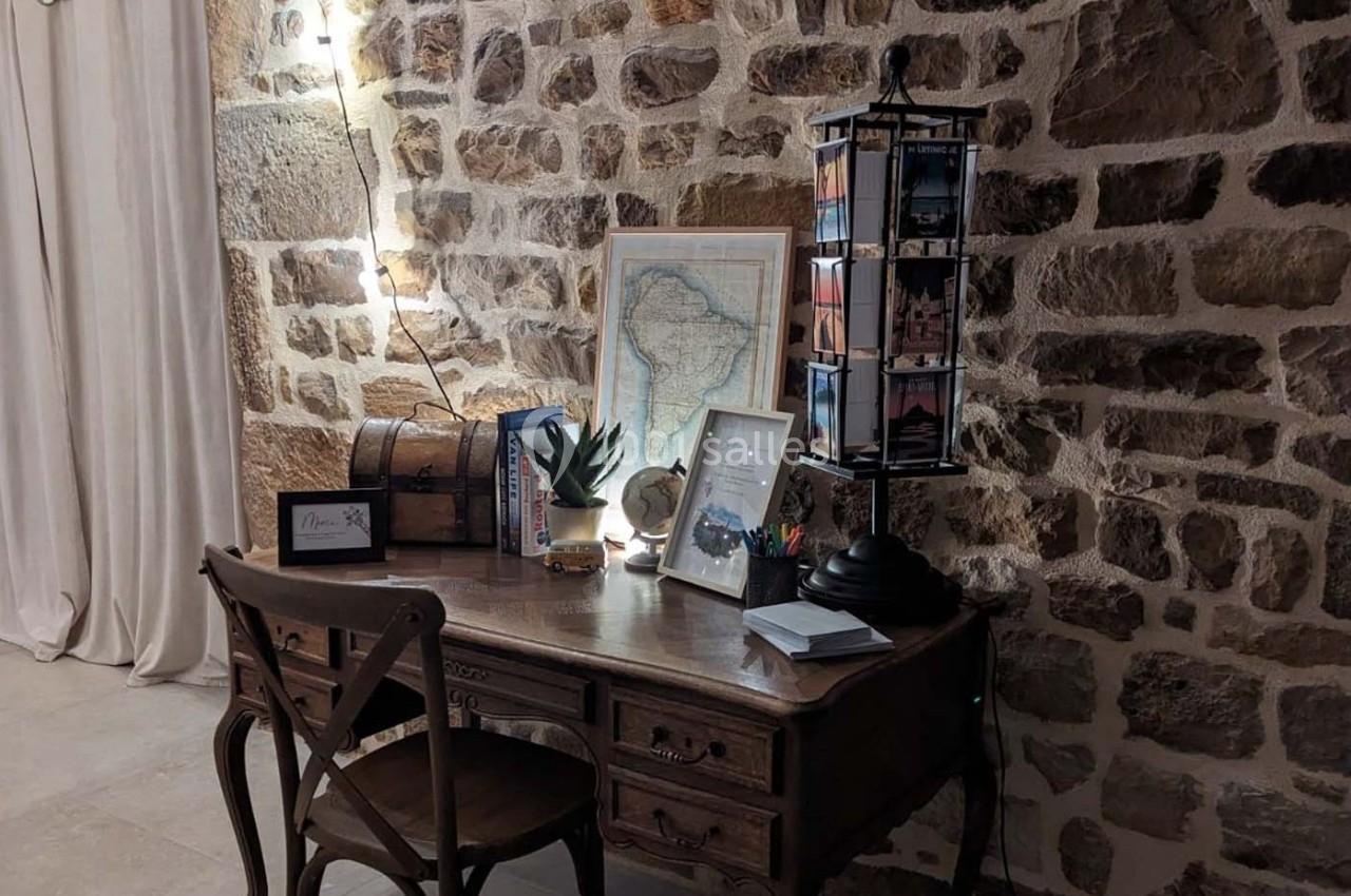 Bureau en bois ancien avec lampe, cartes postales, globe et décoration, devant un mur en pierre apparente.