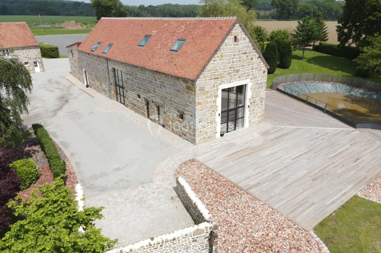 Bâtiment en pierre rénové avec toit en tuiles rouges, entouré d'une terrasse en bois et d'un paysage rural verdoyant.