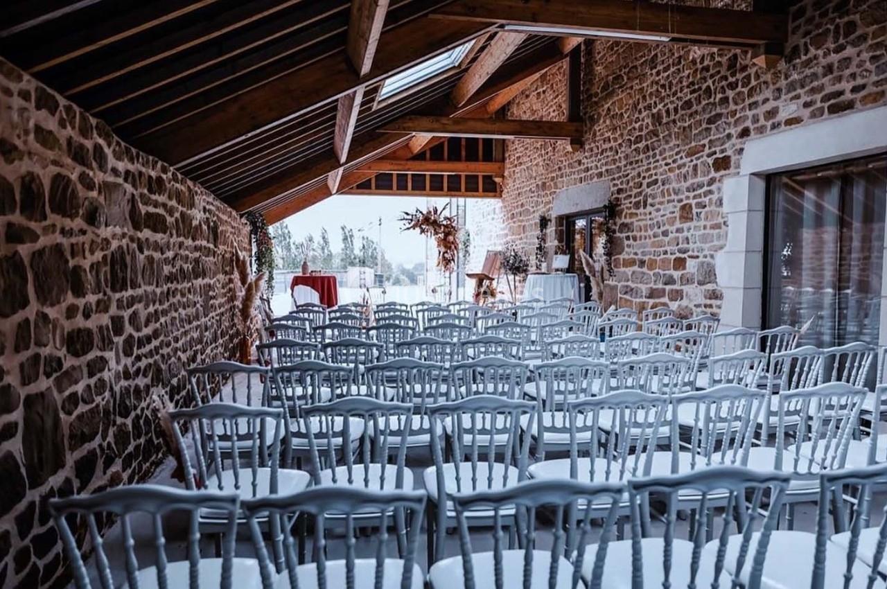 Salle en pierre avec des rangées de chaises blanches alignées, préparée pour un événement avec vue sur l'extérieur.