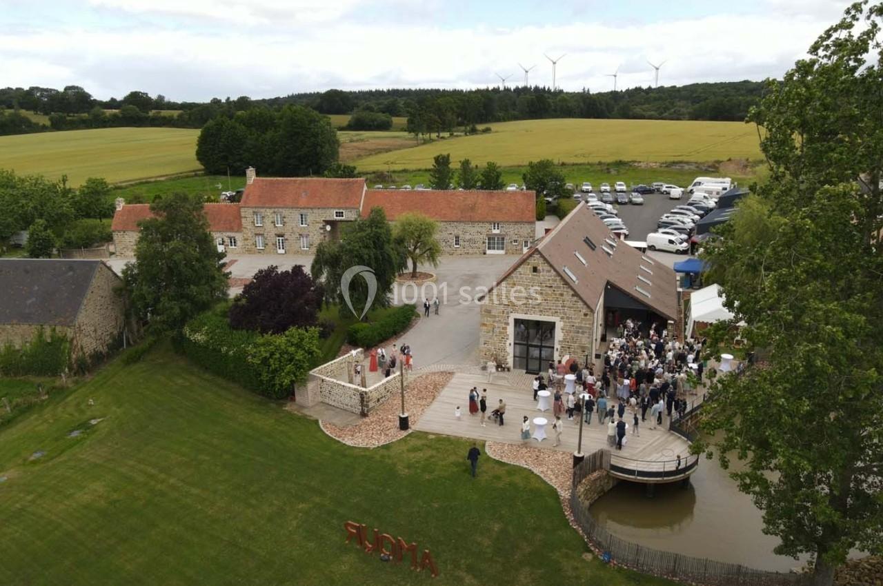 Vue aérienne d'un domaine rural avec bâtiments en pierre, terrasse animée, pelouse et champs environnants.
