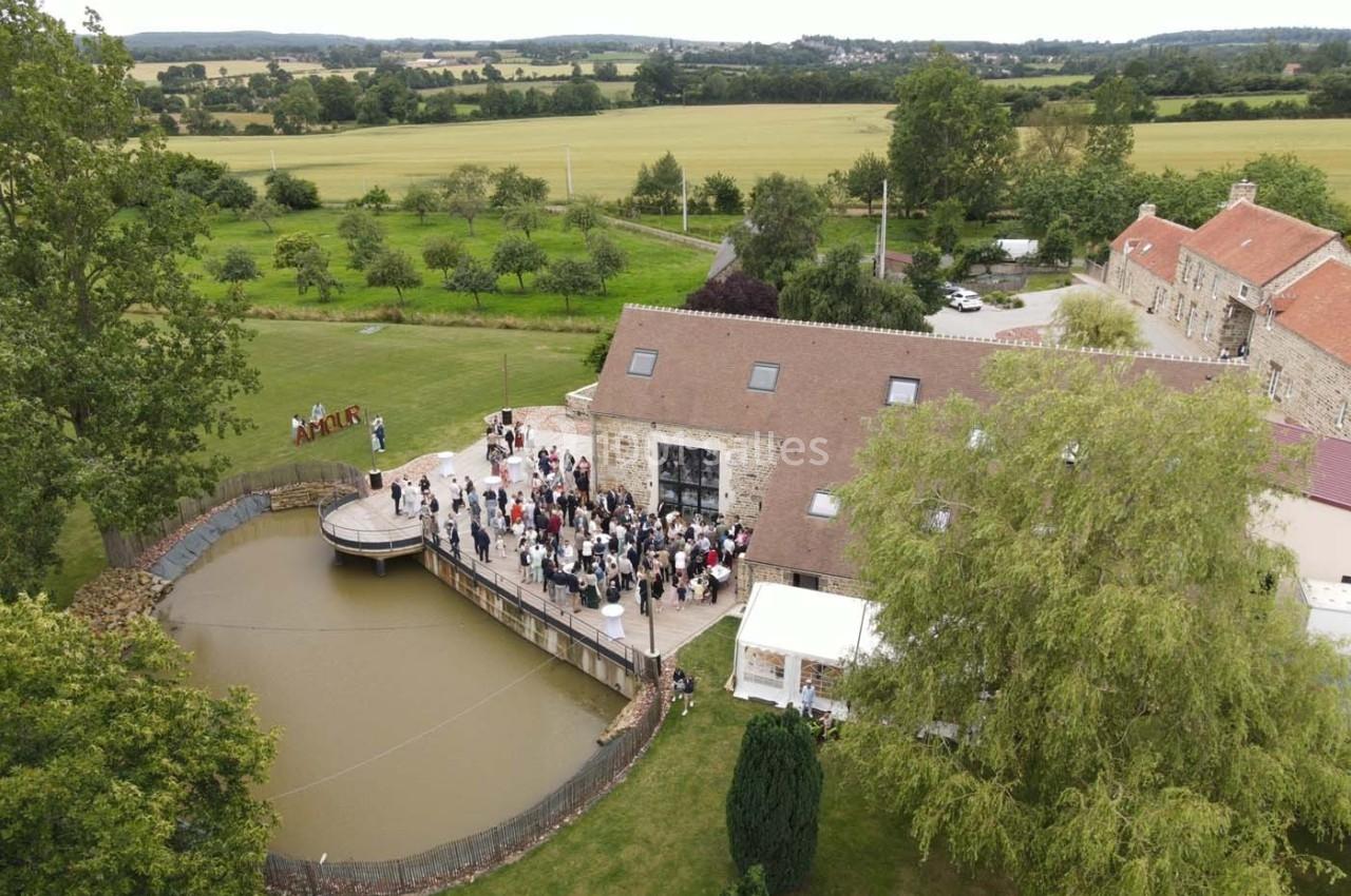 Vue aérienne d'un domaine rural avec un étang, des bâtiments en pierre et un rassemblement de personnes sur une terrasse.