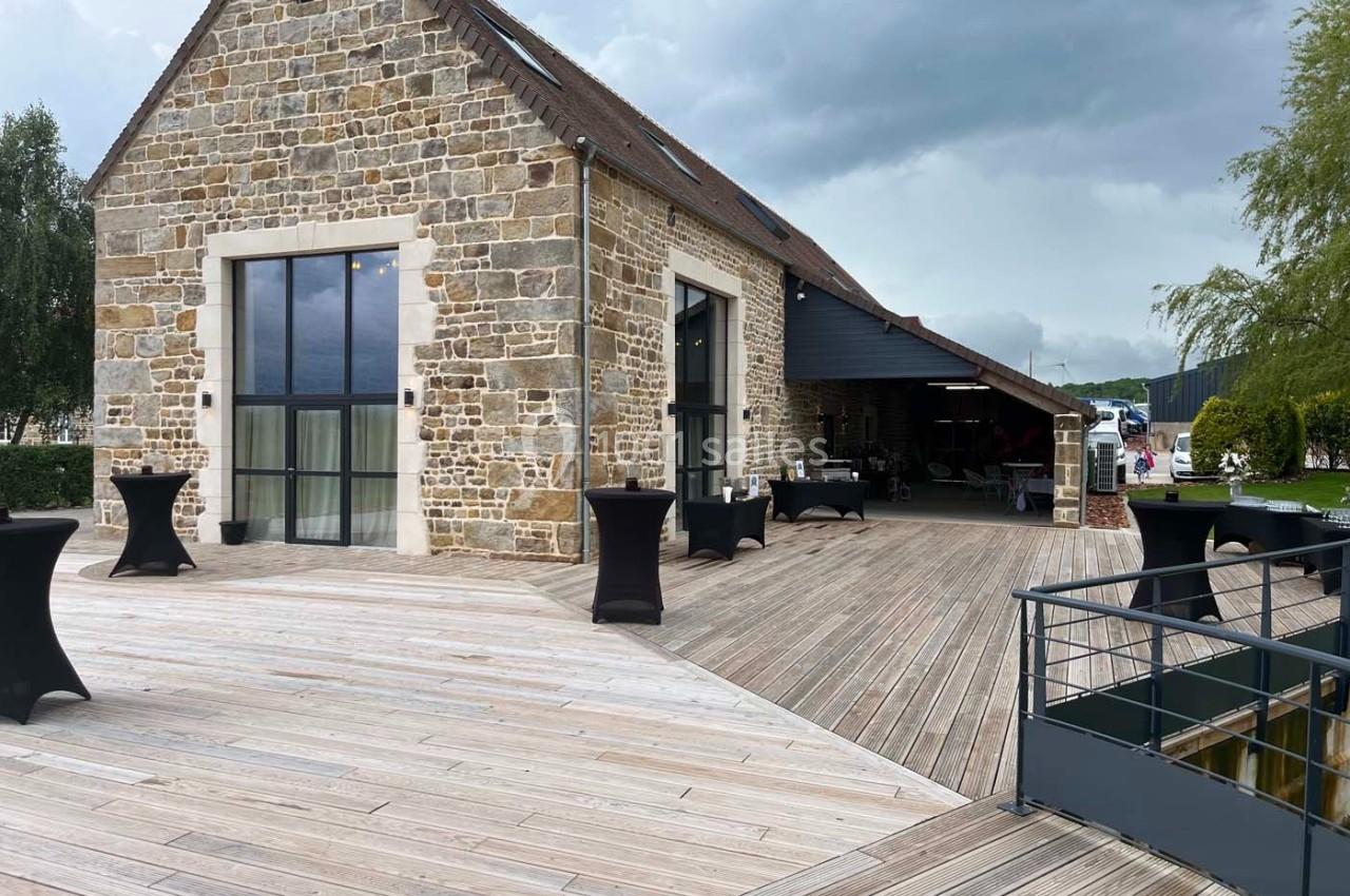 Bâtiment en pierre avec terrasse en bois, grandes baies vitrées et tables hautes, sous un ciel nuageux.
