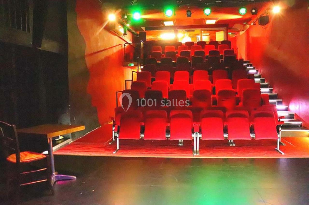 Salle de théâtre avec des rangées de sièges rouges, éclairage coloré et scène au premier plan.
