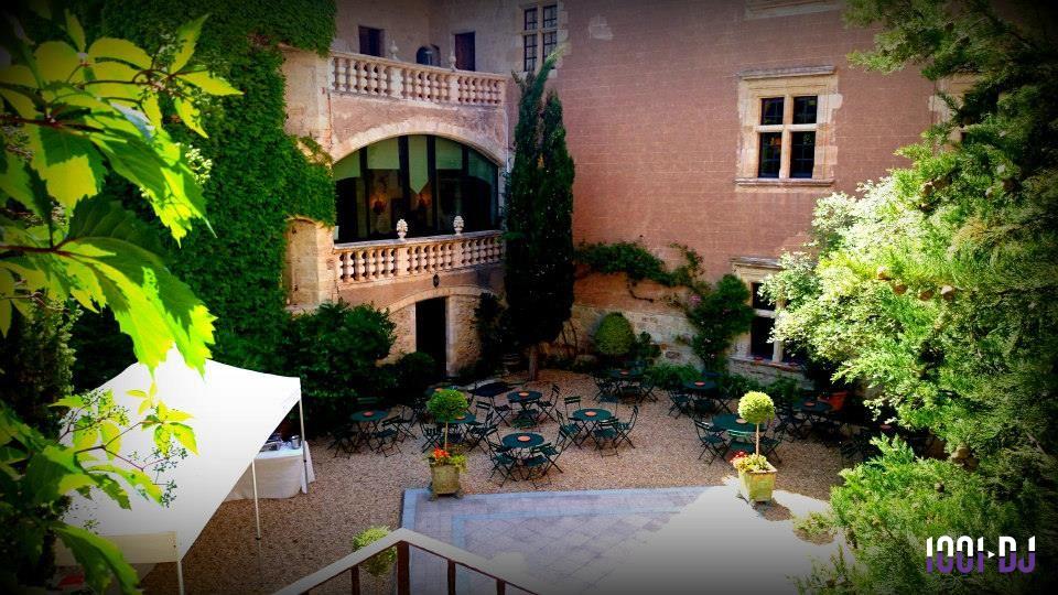 Cour intérieure d'un bâtiment ancien avec terrasse, tables et chaises, entourée de végétation et murs en pierre.