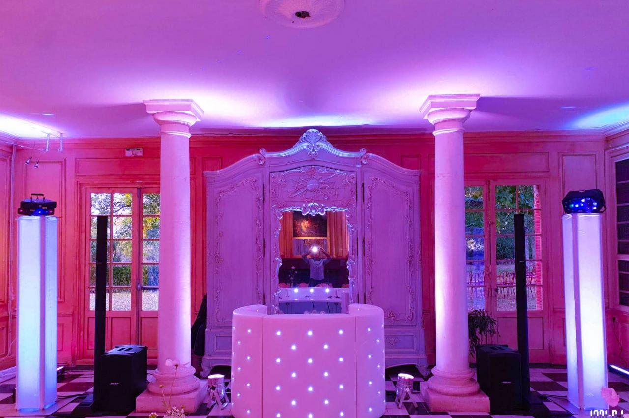 Salle décorée avec des colonnes blanches, un éclairage rose et violet, et une table de DJ lumineuse au centre.