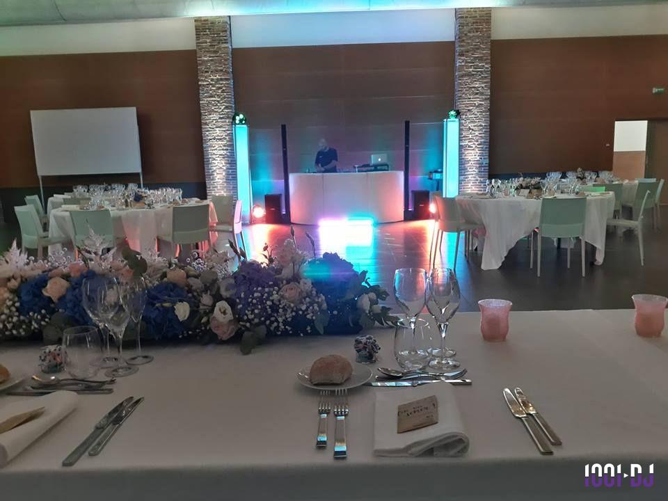 Salle de réception décorée avec des tables dressées, un bouquet floral au premier plan et un DJ en arrière-plan.