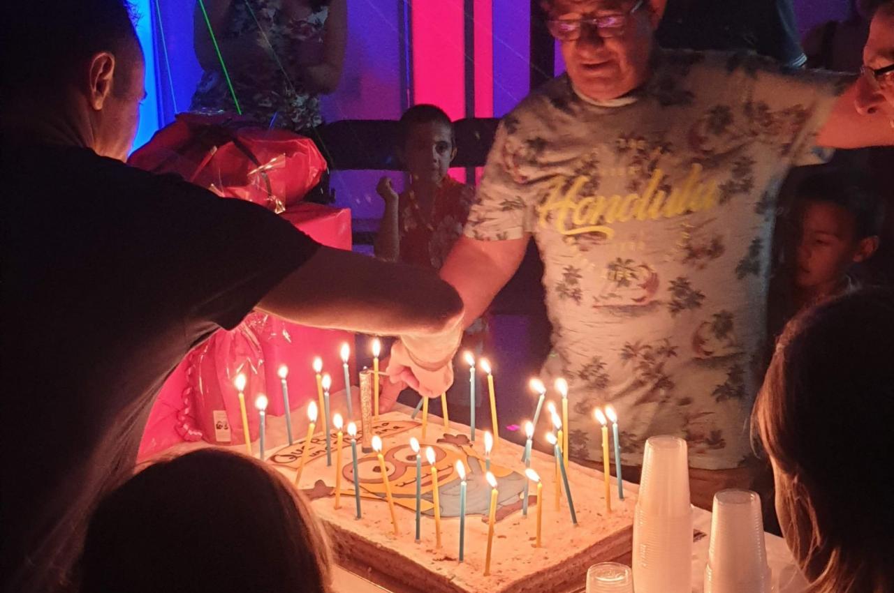 Un homme souffle des bougies sur un grand gâteau d'anniversaire entouré de personnes dans une ambiance festive.