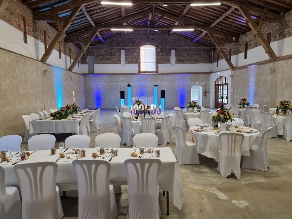 Salle de réception avec tables rondes et rectangulaires décorées, éclairage coloré et murs en pierre apparente.