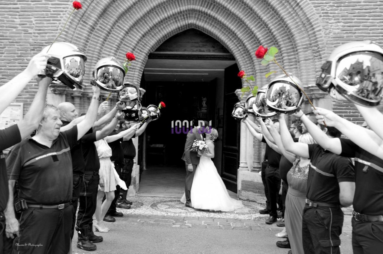 Un couple de mariés s'embrasse sous une haie d'honneur formée par des pompiers tenant des roses rouges.