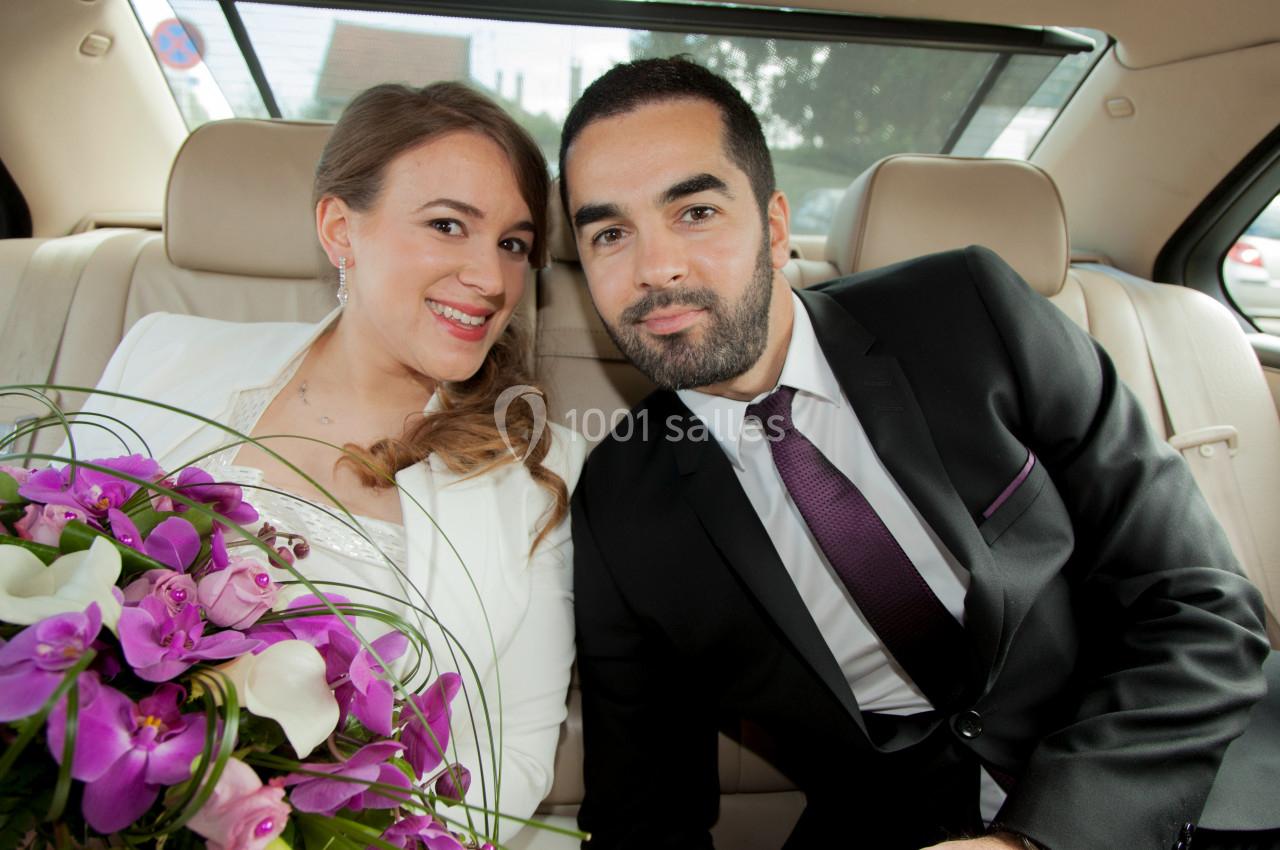 Un couple assis à l'arrière d'une voiture, la femme tenant un bouquet de fleurs violettes et blanches.