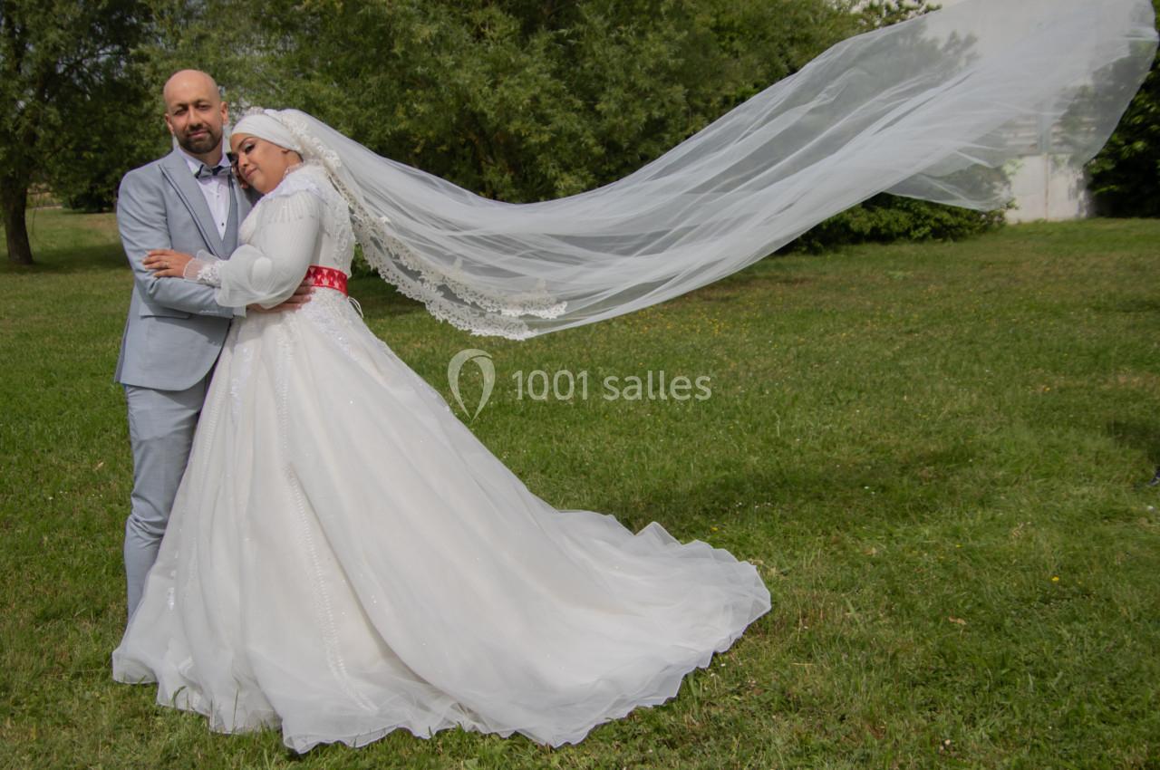 Un couple en tenue de mariage pose dans un parc, la mariée porte un voile long flottant au vent.