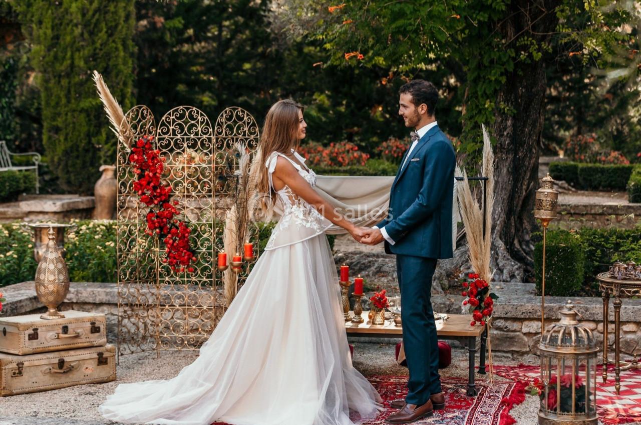 Un couple en tenue de mariage se tient la main dans un décor extérieur orné de fleurs rouges et de lanternes.