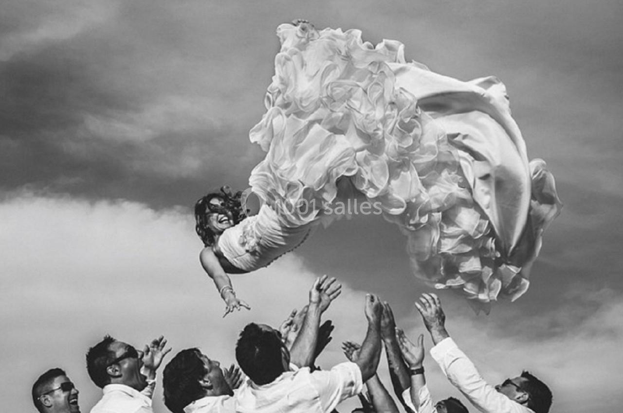 Une mariée en robe blanche est lancée en l'air par un groupe d'hommes vêtus de chemises blanches sous un ciel nuageux.