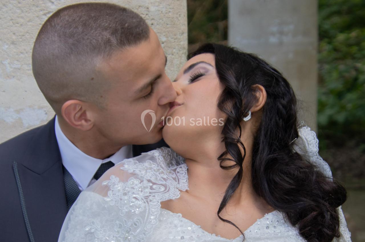 Un couple en tenue de mariage s'embrasse tendrement devant un mur en pierre, entouré de verdure.