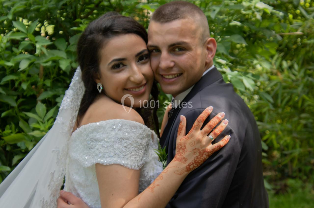 Un couple souriant en tenue de mariage pose devant un fond de verdure.