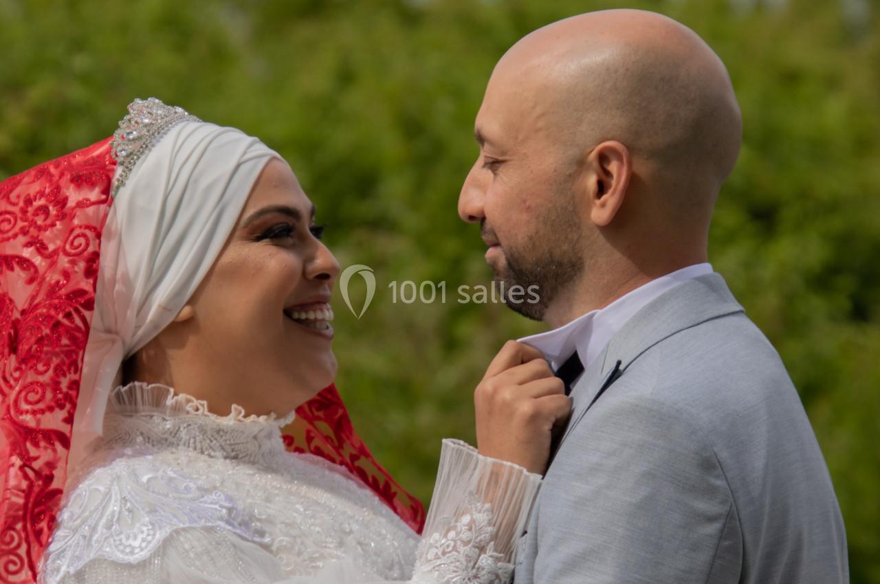 Un couple souriant en tenue de mariage, la mariée ajustant le col de la veste du marié, en extérieur.