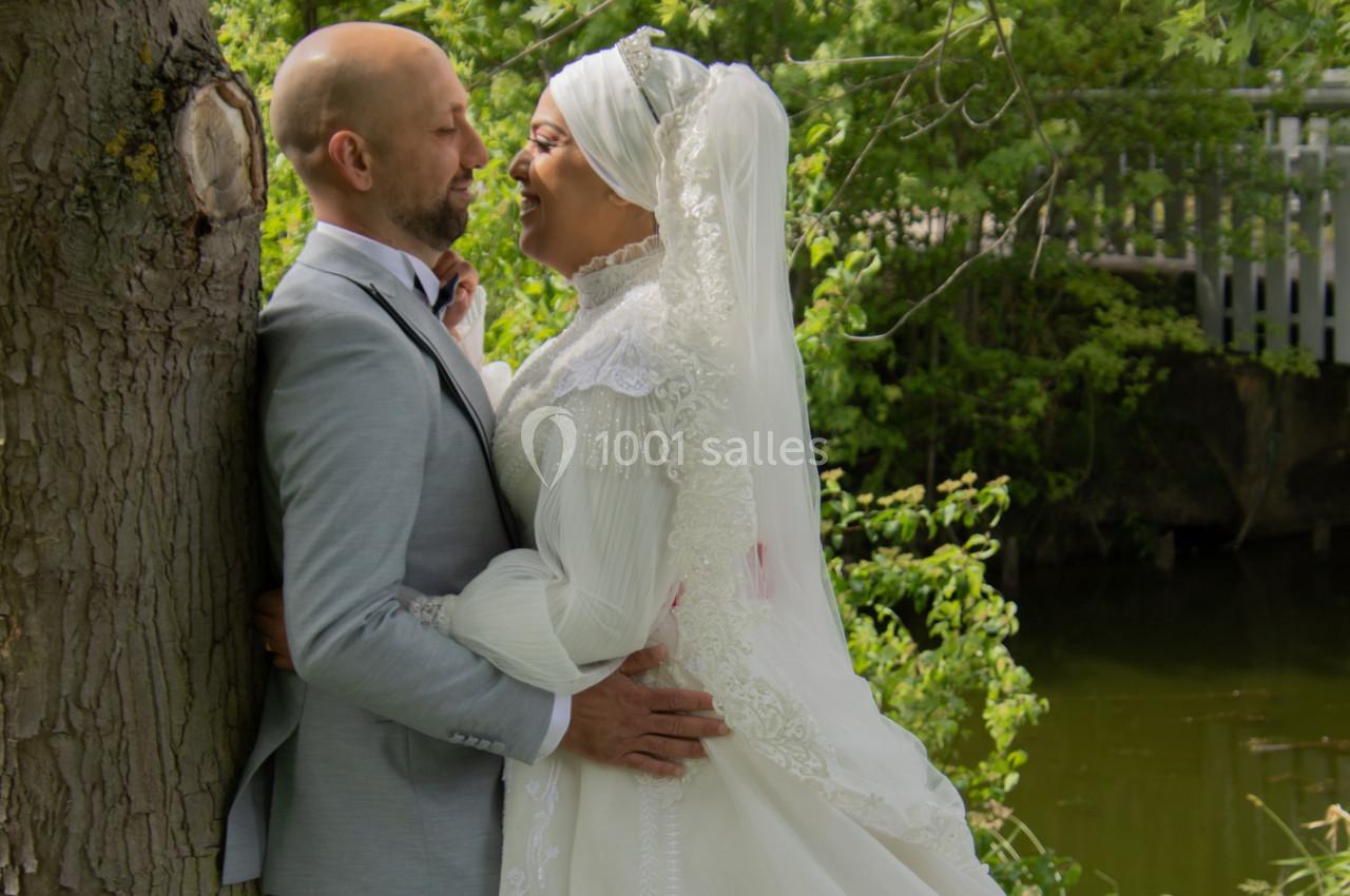Un couple en tenue de mariage se tient près d'un arbre, souriant l'un à l'autre, avec un paysage verdoyant en arrière-plan.