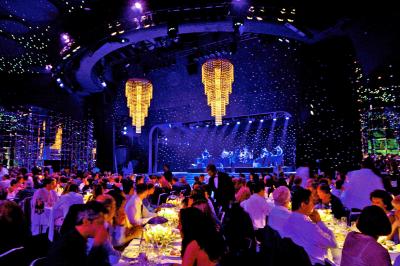 Salle de spectacle illuminée avec des convives dînant devant une scène où des musiciens jouent sous des lustres suspendus.
