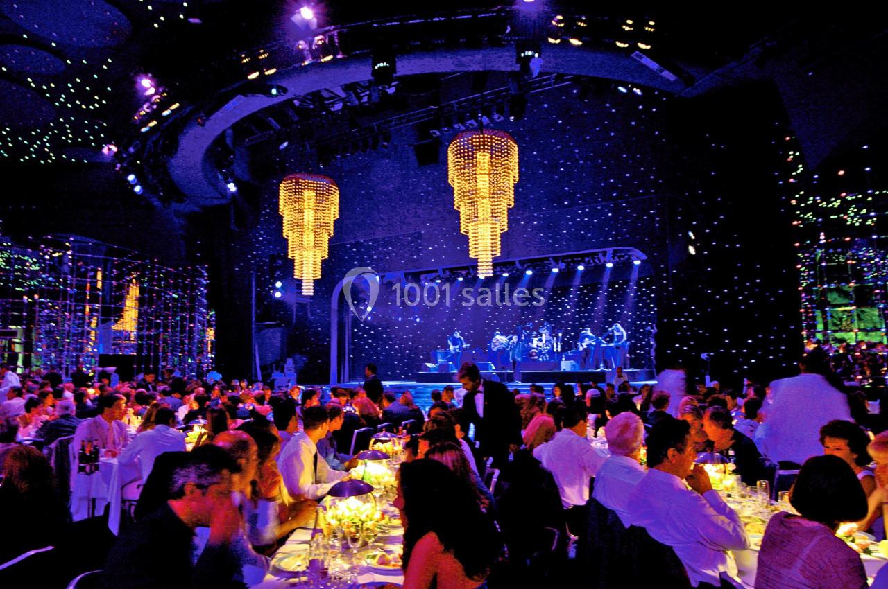Salle de spectacle illuminée avec des convives dînant devant une scène où des musiciens jouent sous des lustres suspendus.
