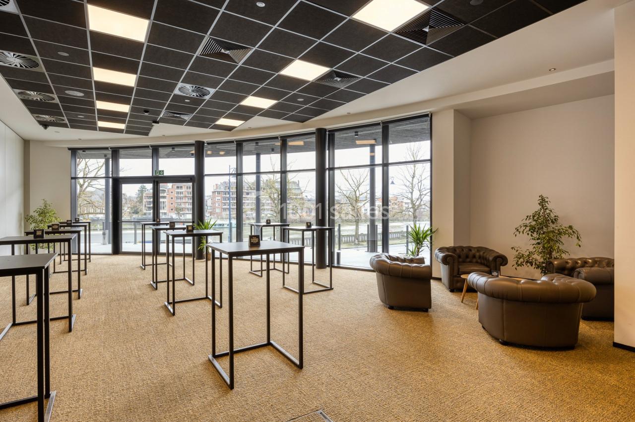 Salle lumineuse avec grandes baies vitrées, tables hautes noires, fauteuils en cuir et plantes décoratives.