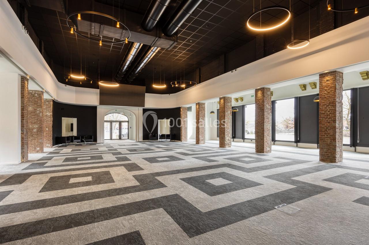 Salle spacieuse avec sol géométrique, colonnes en briques, grandes fenêtres et plafonniers circulaires modernes.