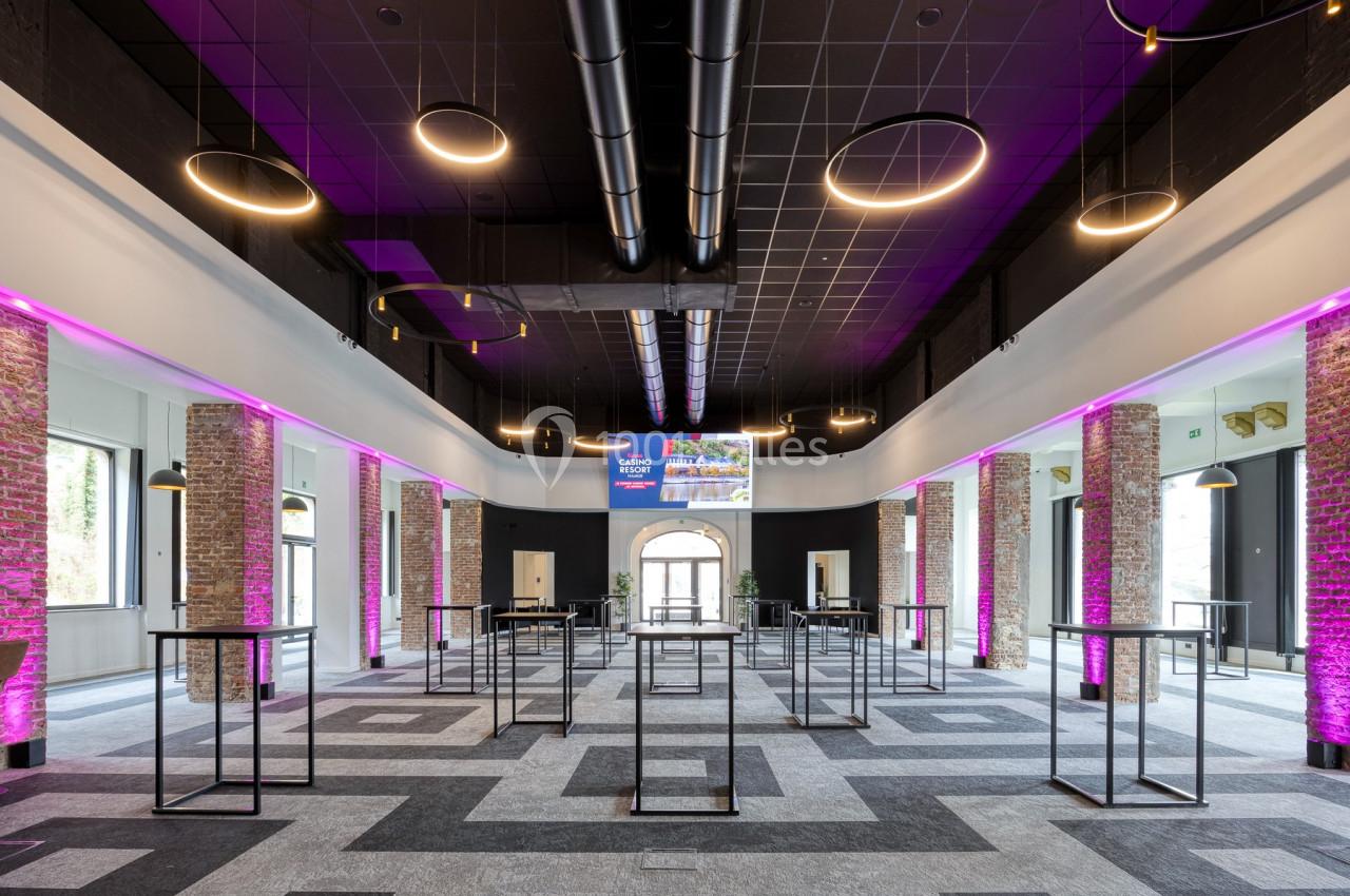 Salle spacieuse avec murs en briques, éclairage moderne, tables hautes et lumière violette accentuant l'ambiance.