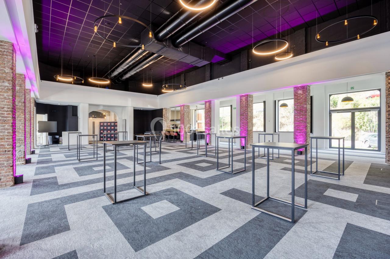 Salle moderne avec un sol à motifs géométriques, des tables hautes métalliques et un éclairage suspendu circulaire.